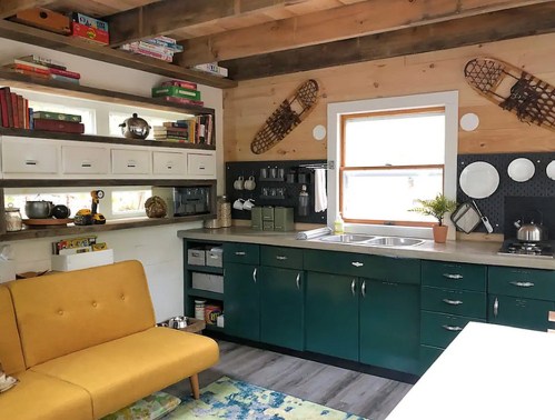 Cozy kitchen with green cabinets, a yellow sofa, bookshelves, and rustic decor, featuring a bright window and modern appliances.