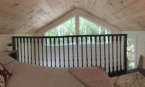 Cozy loft space with wooden walls, a railing, and large windows overlooking a forested area.