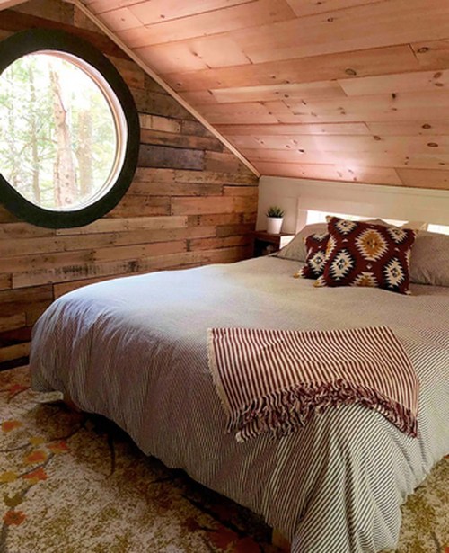 Cozy bedroom with a round window, wooden walls, a patterned blanket, and decorative pillows on a bed.