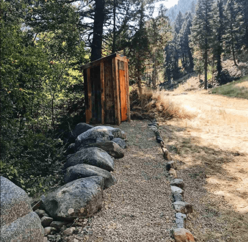 A rustic wooden outhouse beside a gravel path, surrounded by trees and a grassy area in a mountainous landscape.