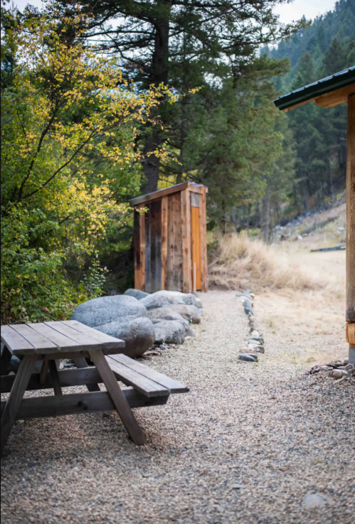 A rustic outdoor scene featuring a wooden picnic table, gravel path, and a small shed surrounded by trees and rocks.