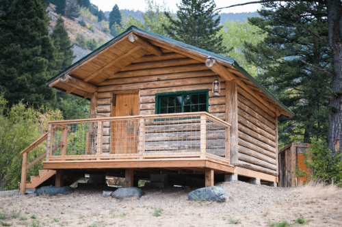 A rustic log cabin with a wooden deck, surrounded by trees and mountains in a natural setting.