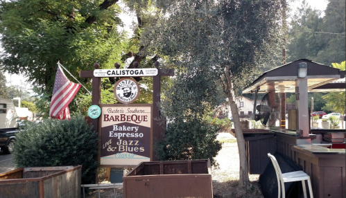 Sign for a barbecue restaurant in Calistoga, featuring bakery, espresso, and live jazz and blues music. American flag nearby.