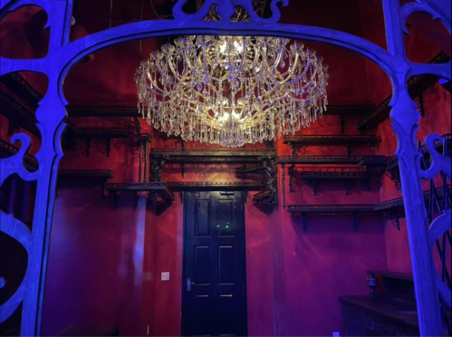 A dimly lit room with red walls, featuring a large chandelier and dark wooden shelves. A blue door is visible.