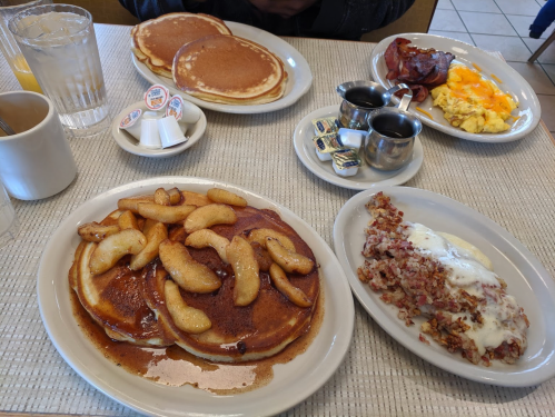 A table set with pancakes topped with apples, sausage, scrambled eggs, bacon, and coffee.