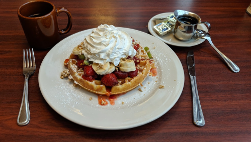 A waffle topped with whipped cream, bananas, strawberries, and syrup, served with a cup of coffee on a wooden table.