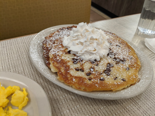 A plate of fluffy pancakes topped with whipped cream and powdered sugar, alongside a serving of scrambled eggs.