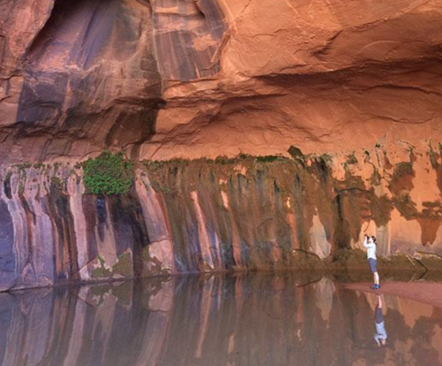 A person stands by a calm water body, reflecting colorful rock formations and a lush green patch on the cliff.