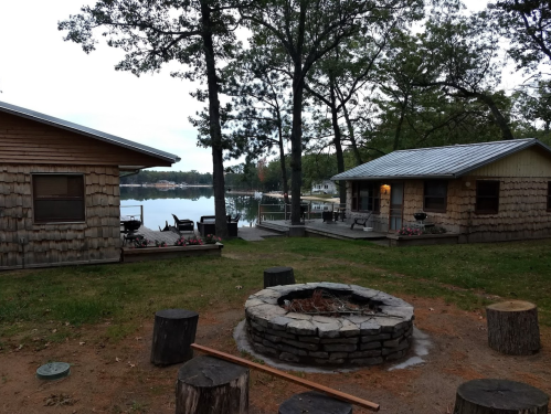Two rustic cabins by a lake, with a stone fire pit in the foreground and trees surrounding the area.