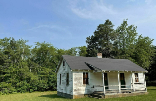 A weathered white house with a black roof, surrounded by green trees and a clear blue sky.