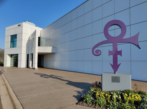 A large purple symbol stands in front of a modern white building, surrounded by colorful flowers.