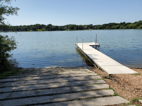 A calm lake with a wooden dock extending into the water, surrounded by greenery and a clear blue sky.