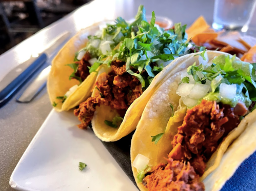 Three tacos filled with seasoned meat, topped with onions and cilantro, served on a plate with tortilla chips.
