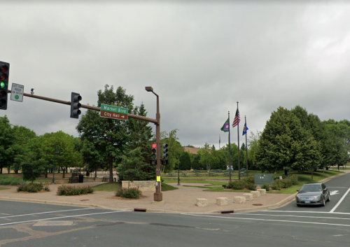 A street view of a city intersection with traffic lights, flags, and a park area near City Hall on a cloudy day.