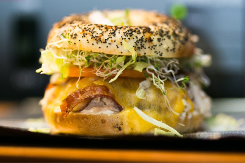 A close-up of a poppy seed bagel sandwich filled with cheese, lettuce, tomato, sprouts, and bacon.