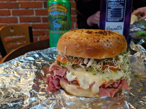A close-up of a sandwich with layers of meat, cheese, sprouts, and tomato, wrapped in foil, next to a can of beer.