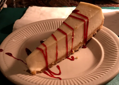 A slice of cheesecake on a plate, drizzled with red sauce, resting on a napkin.
