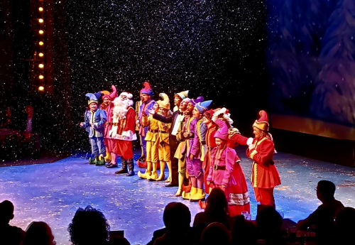 A festive stage with performers in colorful costumes, including Santa, surrounded by falling snowflakes.