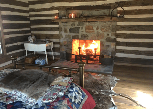 Cozy interior of a rustic cabin with a stone fireplace, quilted bedspread, and warm lighting from candles.