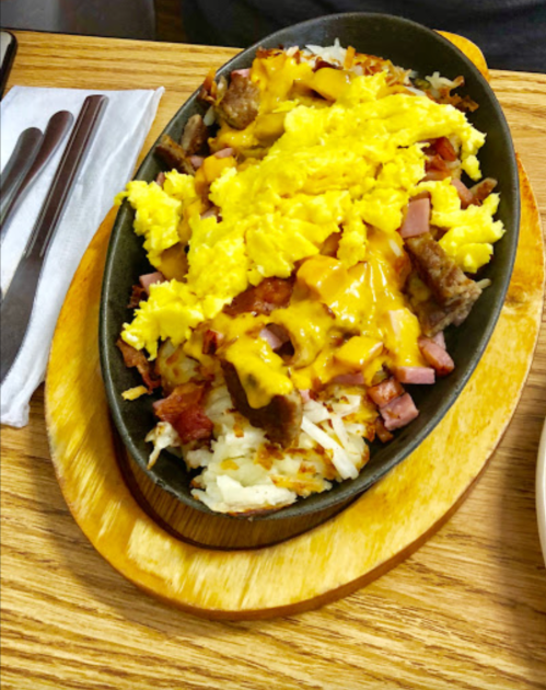 Sizzling platter of rice topped with scrambled eggs, cheese, ham, and beef, served on a wooden board.