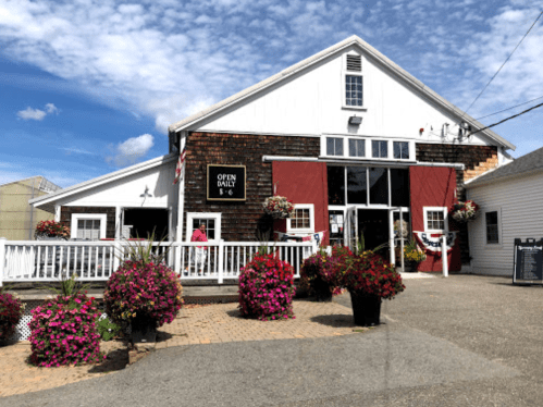 A charming white building with red accents, surrounded by colorful flower pots, under a blue sky. "Open Daily 10-6" sign visible.