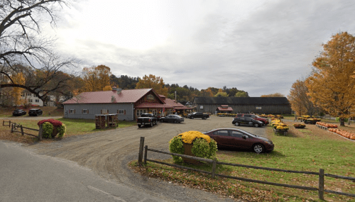 A rustic farm with a red-roofed building, parked cars, and autumn foliage in a rural setting.
