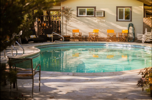 A serene pool area with chairs and a house in the background, surrounded by trees and sunlight.