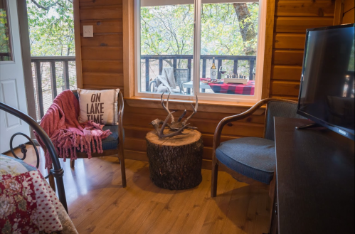 Cozy cabin interior with wooden walls, two chairs, a small table, and a view of trees through a large window.