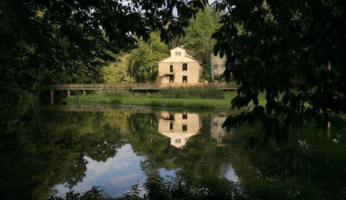 A serene scene of a house reflected in a calm pond, surrounded by lush greenery and trees.