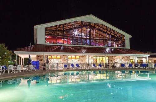 A well-lit building with large windows overlooks a calm pool at night, surrounded by lounge chairs.