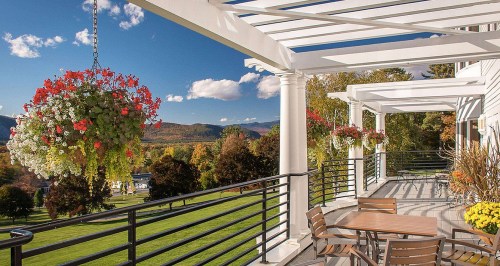 A scenic balcony with hanging flower baskets, overlooking lush greenery and mountains under a clear blue sky.