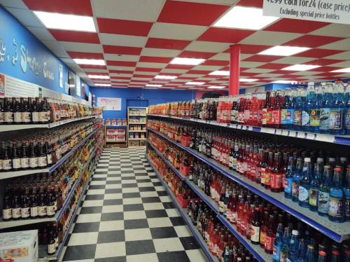 A colorful aisle in a store filled with various soda bottles on shelves, with a checkered floor and red and white ceiling.