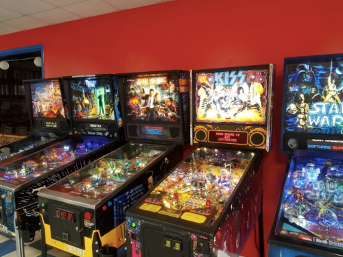 A row of colorful pinball machines featuring various themes against a red wall.