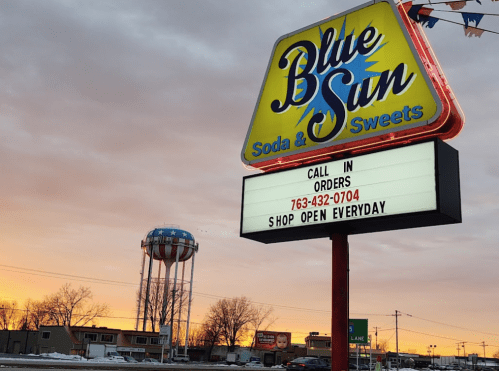 Sign for Blue Sun Soda & Sweets with a colorful logo, water tower in the background, and a sunset sky.