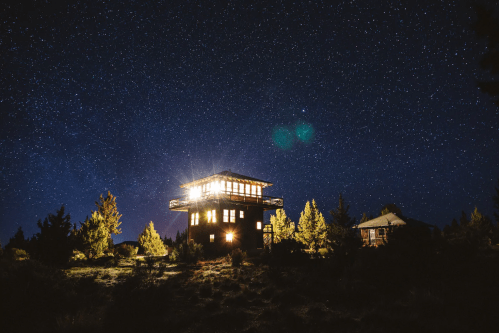 A starry night sky above a rustic cabin with bright lights, surrounded by trees and a serene landscape.