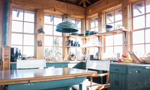 Bright kitchen with wooden beams, large windows, and teal cabinets, featuring a dining table and various kitchenware.