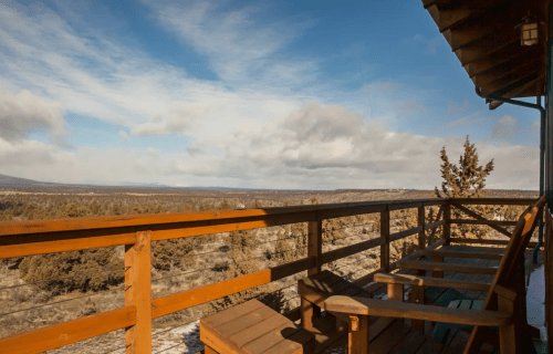 A wooden balcony overlooks a vast landscape of trees and distant mountains under a partly cloudy sky.