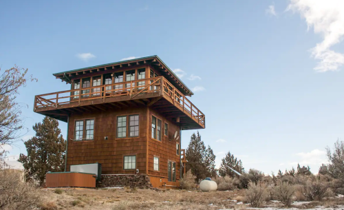 A tall, wooden house with a balcony, surrounded by sparse vegetation and a clear blue sky.