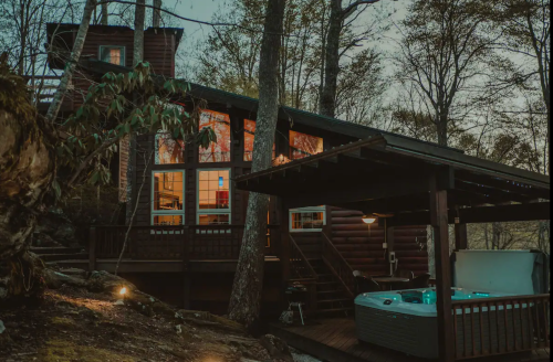 A cozy cabin surrounded by trees, featuring large windows and a hot tub on a deck, illuminated at dusk.
