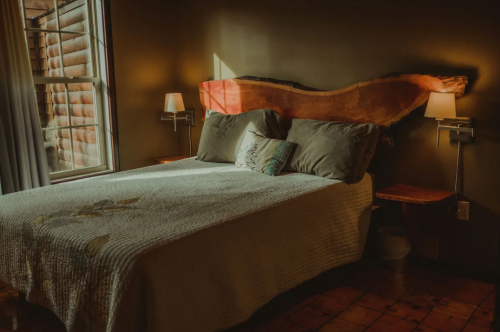 Cozy bedroom with a wooden headboard, soft lighting, and a quilted bedspread. Natural light filters through a window.
