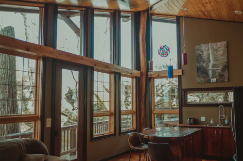 Cozy interior of a modern cabin with large windows, wooden beams, and a view of trees outside.