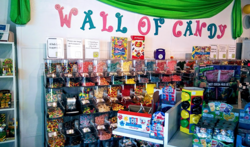 A colorful candy wall filled with various jars of sweets and treats, labeled "WALL OF CANDY" above.