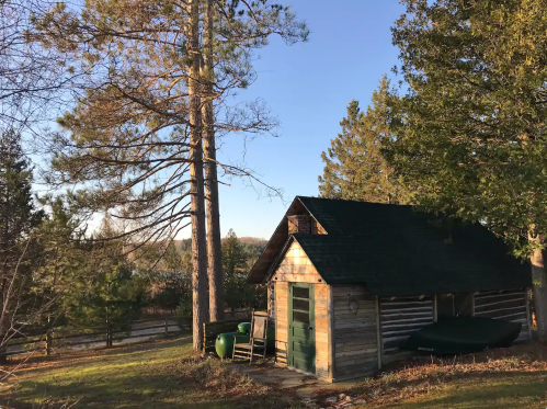 A rustic wooden cabin surrounded by tall trees and a clear blue sky, set in a peaceful natural landscape.