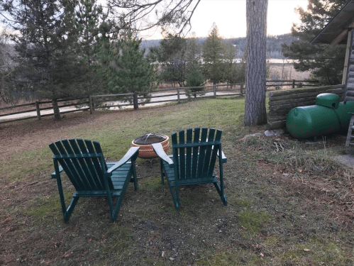 Two green adirondack chairs facing a fire pit, surrounded by trees and a grassy area, with a green tank nearby.