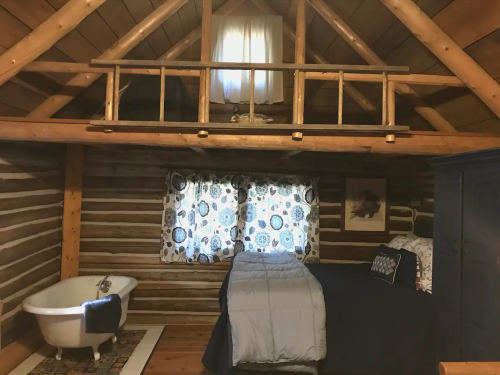 Cozy log cabin interior featuring a bed, bathtub, and wooden beams, with patterned curtains and a loft area above.