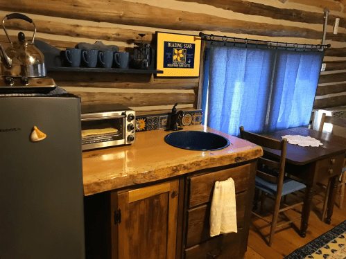 Cozy rustic kitchen with wooden walls, a blue sink, small appliances, and a dining table by a window with blue curtains.