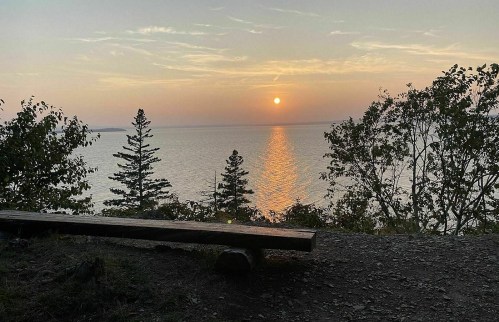 A serene sunset over a calm lake, with trees framing the view and a wooden bench in the foreground.