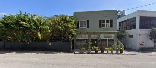 A two-story building with green walls and large windows, surrounded by palm trees and potted plants on a sunny street.