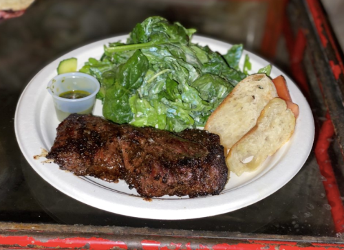 A plate with grilled meat, fresh greens, and slices of bread, accompanied by a small container of sauce.