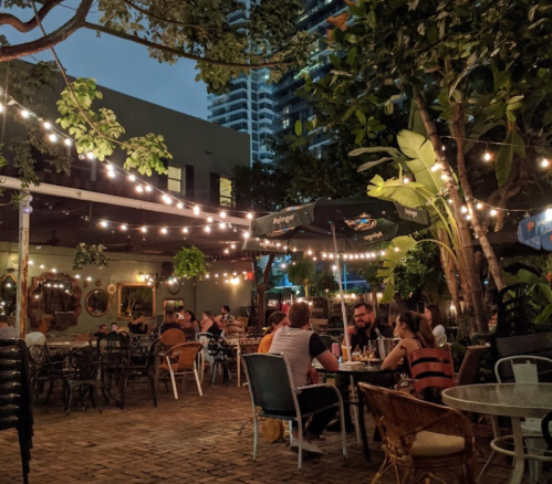 A cozy outdoor dining area with string lights, plants, and people enjoying meals at tables in the evening.